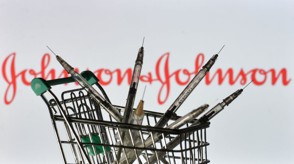 Medical syringes in mini shopping trolley are seen in front of the Johnson and Johnson logo displayed on screen.